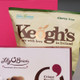 Close up. A partially visible bag of Keogh's snacks features the text "Gluten free" and "Grown with love in Ireland." Below it, a box of Lily O'Brien's chocolates showcases the label "Crispy Hearts" along with a tagline about being passionate about chocolate. The background is a vibrant pink. Close up. A partially visible bag of Keogh's snacks features the text "Gluten free" and "Grown with love in Ireland." Below it, a box of Lily O'Brien's chocolates showcases the label "Crispy Hearts" along with a tagline about being passionate about chocolate. The background is a vibrant pink.